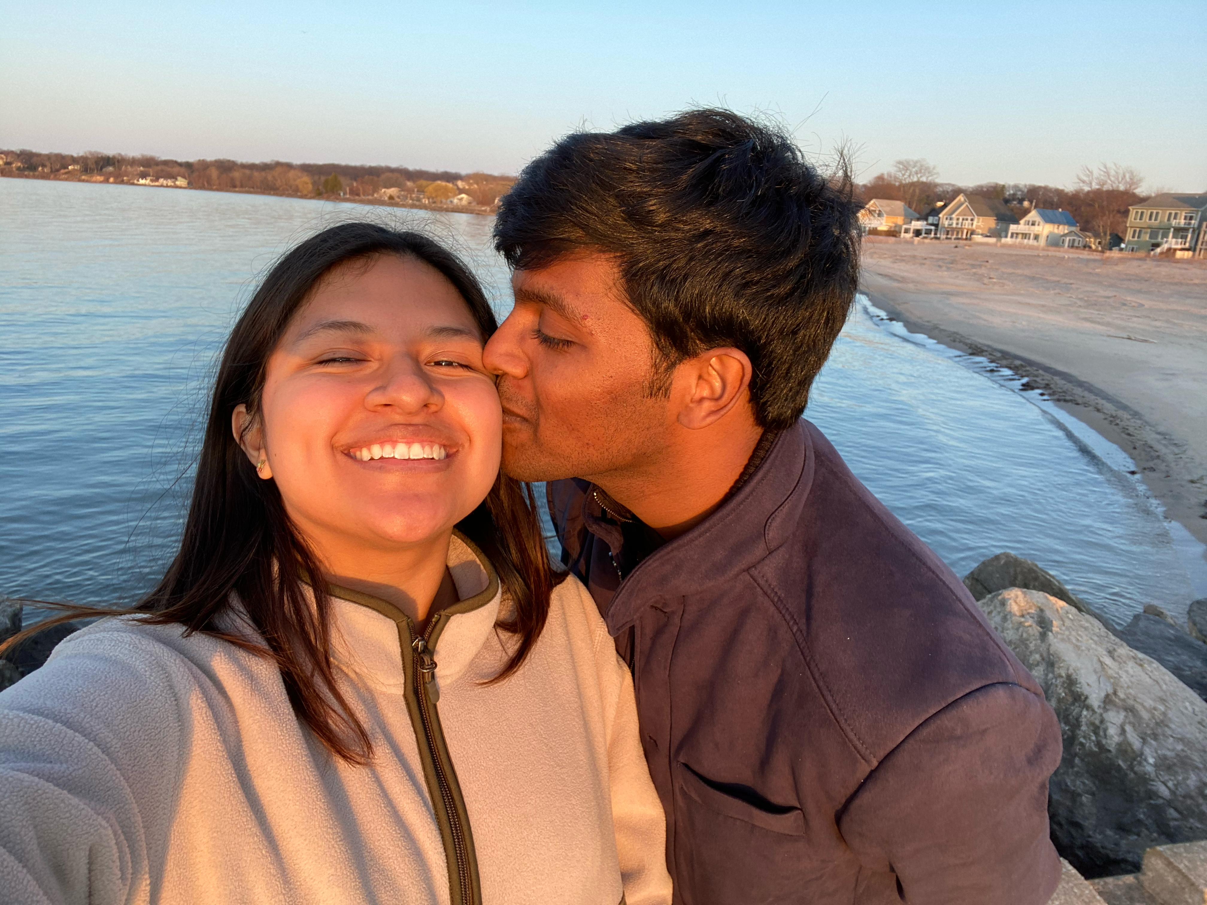 Aravind kissing Tiffany on the cheek at sunset by the beach